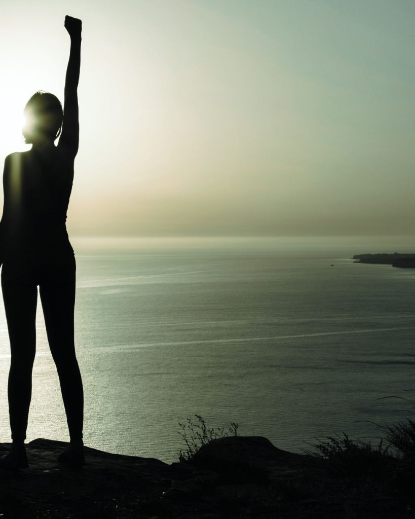Silhouette of woman stretching with one arm raised as the sun rises over the water.